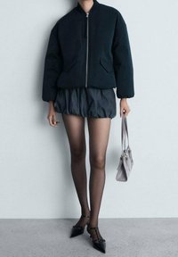 Giubbotto bomber nero con zip frontale, abbinato a una gonna scura a balze, collant neri trasparenti e scarpe nere a punta. Tiene una borsa grigio chiaro.
