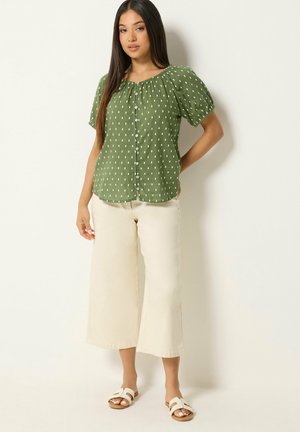 Mujer con pelo largo y oscuro que lleva una blusa verde de manga corta con botones y puntos blancos, pantalones crema de pierna ancha y corte cropped, y sandalias blancas.