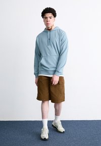 Sudadera azul claro con bolsillo tipo canguro, pantalones cortos de cargo marrones, calcetines blancos y zapatillas gris-verde. Conjunto simple y casual.