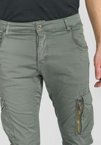 Groene cargobroek gemaakt van lichtgewicht stof, voorzien van een ritsvak aan de zijkant, knoopsluiting en steekzakken aan de voorkant.