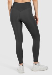 Leggings noirs taille haute en matériau extensible, avec une texture lisse et une coupe ajustée, associés à des chaussures de sport blanches.