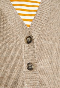 Beige stickad kofta med V-ringning, ribbade kanter och två mörka knappar. En randig orange och vit skjorta syns under.