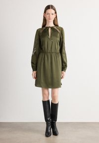 Olijfgroene jurk met lange, doorzichtige kantmouwen, een striksluiting bij de hals en een elastische taille. Gepaard met zwart, kniehoog laarzen.