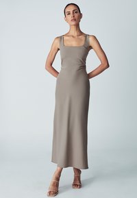 Langes, figurbetontes Kleid in einem hellen Taupe mit quadratischem Ausschnitt und breiten Trägern. Glatte, dehnbare Stoff; minimale Verzierungen.