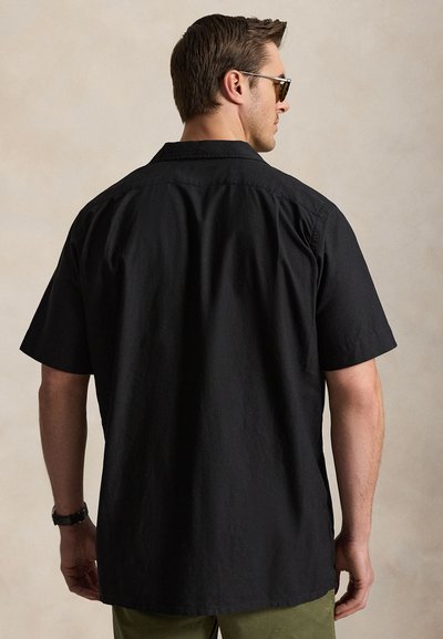 Man draagt een zwart overhemd met korte mouwen en knopen, een zonnebril en een polshorloge, met zijn rug naar de kijker gekeerd tegen een effen beige achtergrond.