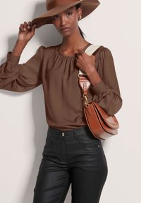 Braune Bluse mit langen Ärmeln und gerafftem Ausschnitt, kombiniert mit schwarzen, schmalen Hosen. Eine braune Leder-Handtasche mit einem beigen Riemen rundet den Look ab.