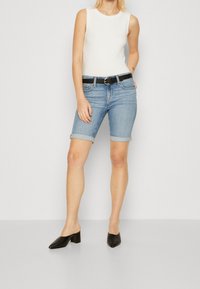 Vaaleansiniset farkkushortsit käännetyillä lahkeilla, yhdistettynä valkoiseen hihattomaan toppiin ja mustaan vyöhön. Päällä mustat korolliset kengät.