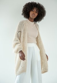 Beżowy sweter z dzianiny z otwartym przodem, charakteryzujący się luźnym krojem i skomplikowanymi wzorami koronkowymi. Nosi się go na jasnobeżową bluzkę i białe spodnie.