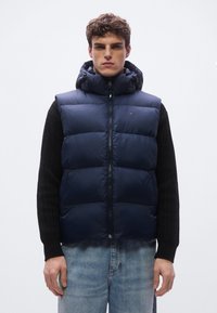 Colete puff azul-marinho com capuz ajustável, design acolchoado, fecho frontal de correr. Combinado com um suéter preto texturizado e jeans azul claro.