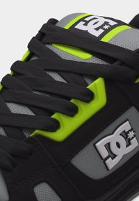 Κοντινή λήψη μαύρου και γκρι αθλητικού παπουτσιού με μαύρα κορδόνια και φωσφορίζουσες κίτρινες τρύπες για τα κορδόνια, με λευκό λογότυπο DC Shoes στη γλώσσα και στη πλευρά.