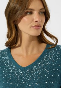 Maglione teal con scollatura a V, caratterizzato da un tessuto testurizzato e da strass sparsi sul davanti, che creano un effetto scintillante.