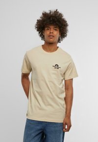 Beige cotton t-shirt with short sleeves, featuring a black graphic text and logo on the left chest. Model stands in casual blue jeans.