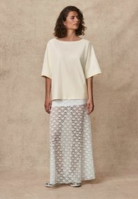 Světlounce žluté oversized bavlněné tričko s širokými rukávy, spárované s dlouhou bílou krajkovou sukní se květinovými vzory a průsvitnou texturou.