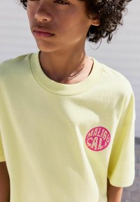 Lichtgele katoenen t-shirt met een roze grafisch logo aan de linkerkant. Korte mouwen en een ronde hals. Gladde textuur.