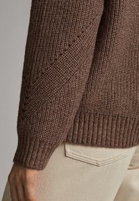 Maglione marrone lavorato a maglia con un motivo testurizzato, polsini a coste e orlo aderente. Indossato con pantaloni beige chiaro, in mostra il dettaglio della cucitura laterale.