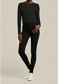 Personne portant un haut noir ajusté à manches longues et un jean skinny noir, debout avec une main dans la poche, baskets blanches visibles.