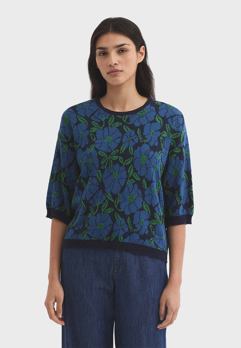 Frau trägt einen hellblauen Pullover mit kurzärmeligem Floralmuster und dunkelblaue Jeans, steht vor einem schlichten hellen Hintergrund.