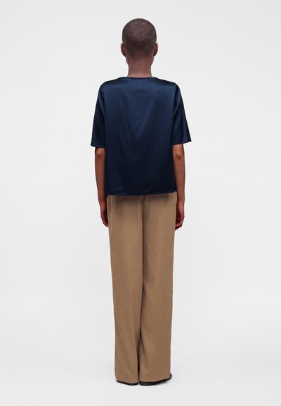 Personne debout de dos portant une chemise bleu marine à manches courtes et un pantalon beige à jambes larges sur un fond uni.