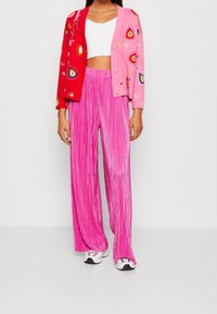 Cardigan tricoté avec des sections rouges et roses, présentant des motifs colorés et des boutons, associé à un pantalon large plissé rose et des baskets.