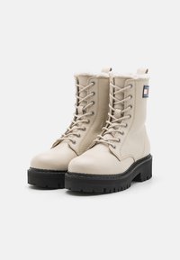 Botas beige de cuero con cordones, forro acolchado, puntera redonda, suela negra gruesa y parche con logo en el costado.