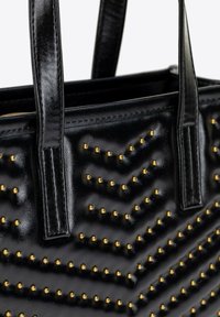 Černá kožená taška typu tote s chevronovým vzorem, zdobená zlatými cvočky pro texturu. Disponuje pevnými, plochými uchy a zipem na vrchu.