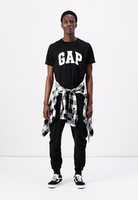 Black t-shirt with "GAP" logo, black pants, and black-and-white plaid shirt tied at the waist. Black and white sneakers complete the look.