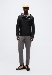 Άνδρας που στέκεται φορώντας μπεζ καπέλο, μαύρο μπουφάν με φερμουάρ, γκρι παντελόνι και μαύρα και μπεζ παπούτσια πεζοπορίας σε λευκό φόντο.