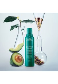 Aveda botanisk reparations stärkande schampo i en tealflaska, med skivad avokado och glasbehållare som visar frön och blad.
