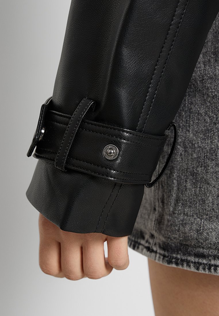 Schwarze Lederjackenmanschette mit glatter Oberfläche und einem Riemen-Detail, gesichert durch einen silbernen Druckknopf. Sichtbare Hand mit heller Hautfarbe.