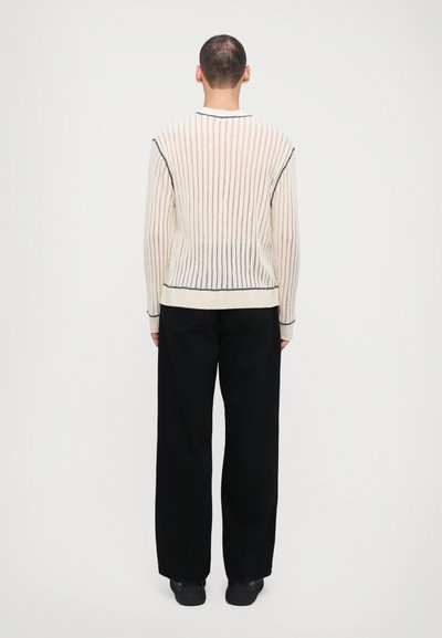 Cream ribbed knit sweater with navy accents, featuring long sleeves and a fitted silhouette, paired with loose black trousers and dark footwear.