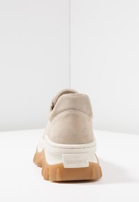 Beiger Wildledersneaker mit einer klobigen weißen Zwischensohle und braunem Gummiaußensohle. Er verfügt über eine abgerundete Rückseite und ein eingeprägtes "BRONX"-Logo.