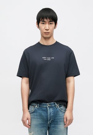 Tricou din bumbac albastru închis, cu mâneci scurte, având text alb „Good Times and Tan Lines” centrat pe piept, asortat cu blugi albaștri cu aspect uzat.