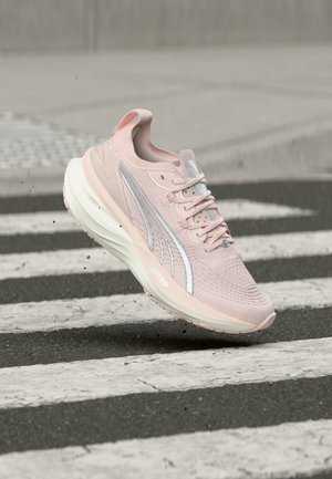 Světle růžová běžecká bota s bílou podrážkou a stříbrným logem, zachycena ve vzduchu nad černobílým pruhovaným povrchem.