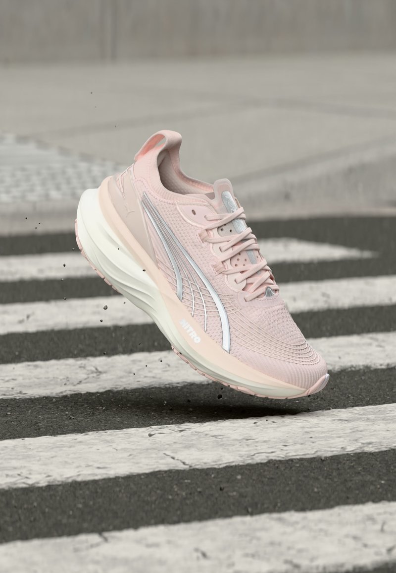 Ανοιχτό ροζ αθλητικό παπούτσι με λευκή σόλα και ασημί λογότυπο, αιωρείται στον αέρα πάνω από μαύρο και άσπρο ριγέ πεζοδρόμιο.