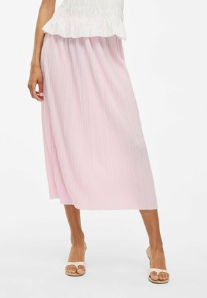 Jupe plissée rose clair avec un design à mi-longueur, présentant une texture lisse et un subtil éclat, associée à des sandales de ton neutre.