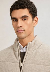 Junger Mann mit lockigem, dunklem Haar trägt einen beigen Reißverschluss-Pullover über einem weißen Hemd mit Kragen vor neutralem Hintergrund.