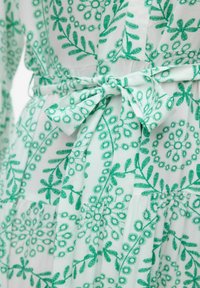 Grön blommönstrad klänning i tyg med knytning vid midjan, i en ljus bakgrund med texturerade blad- och blomdesigns.