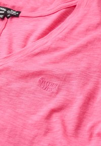 Camiseta rosa de manga corta con escote en V, hecha de un tejido suave y ligero. Presenta un sutil logo en la esquina inferior izquierda. Acabado texturizado.