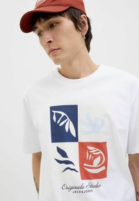 Hvid t-shirt med et multifarvet grafik med blå, røde og cremefarvede firkanter samt bladmønstre. Materialet virker let og afslappet.