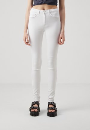 Femme portant un jean skinny blanc et des sandales noires épaisses avec des boucles, debout devant un fond clair uni.