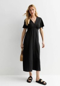 Vestido largo negro con mangas cortas, escote en V y detalle de botones en el frente; combinado con sandalias de tiras negras texturizadas y un bolso de mimbre.