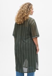 Khaki-Mesh-Kimono mit kurzen Ärmeln, das ein gestreiftes Muster, Seitenschlitze und kontrastierende Bündchen aufweist. Getragen über hellblauen Jeans.