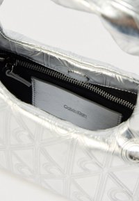 Ασημένια τσάντα με ανάγλυφο μοτίβο, με μερικώς ανοιχτό φερμουάρ που αποκαλύπτει ένα ασημένιο πορτοφόλι Calvin Klein στο εσωτερικό.