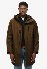 Brązowa wodoodporna kurtka z kapturem, czarnymi akcentami, przednią kieszenią na zamek i luźnym krojem, noszona na czarnym swetrze i niebieskich dżinsach.