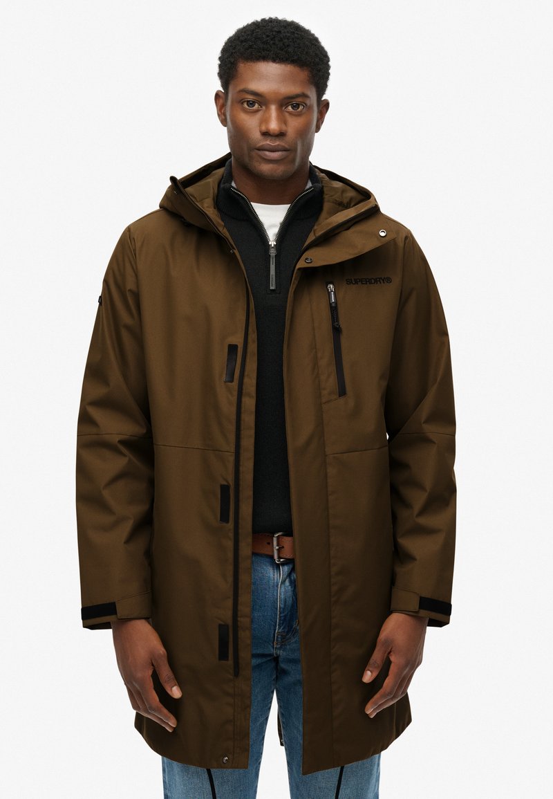 Brązowa wodoodporna kurtka z kapturem, czarnymi akcentami, przednią kieszenią na zamek i luźnym krojem, noszona na czarnym swetrze i niebieskich dżinsach.