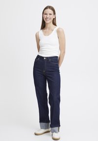 Weißes ärmelloses Tanktop, dunkelblaue hoch taillierte Jeans mit sichtbaren Nähten und aufgerollten Säumen, kombiniert mit hellen Sneakers.