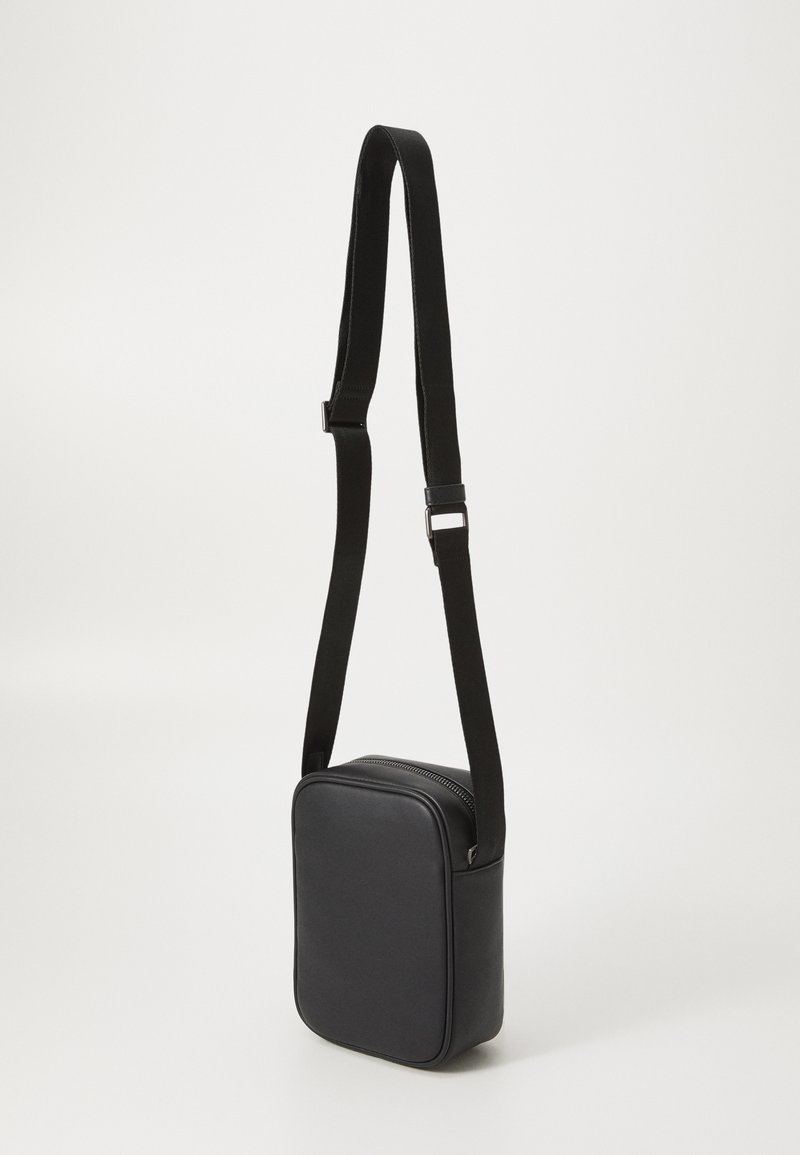 Bolso bandolera de imitación cuero negro con forma rectangular, cierre con cremallera y correa ajustable, que presenta una textura suave y un diseño minimalista.