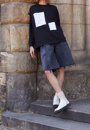 Zwart shirt met lange mouwen met witte grafische patches, gecombineerd met losse grijze denim korte broeken met rafelige randen. Witte open-toe schoenen met zwarte zool.