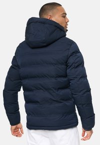 Marinblå pufferjacka med huva, tillverkad av quiltat material. Har en strömlinjeformad silhuett och ribbad mudd för värme och komfort.
