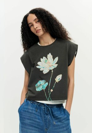 Kobieta o kręconych włosach, ubrana w czarny bezrękawnik z wielokolorowym kwiatowym wzorem oraz niebieskie spodnie ze ściągaczem, z rękami w kieszeniach.